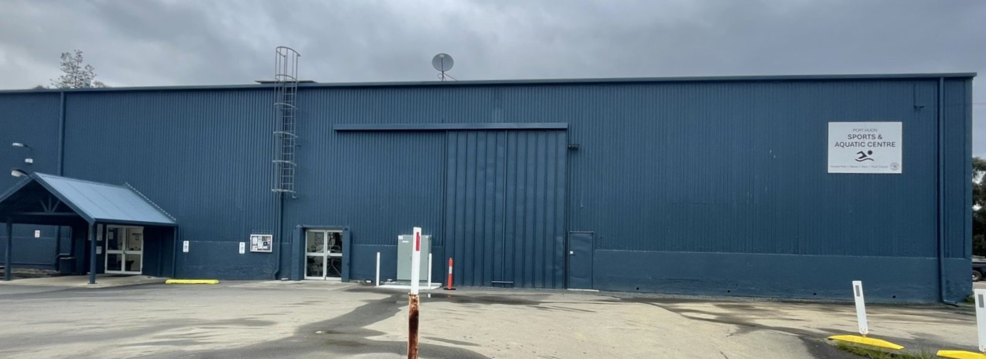 A large blue metal building with a small covered entrance on the left, a wide central garage door, and a sign reading “Sports Aquatic Centre” on the right under a cloudy sky. The ground is wet and mostly empty.