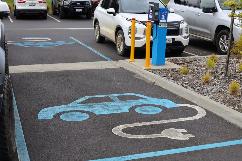 Electric vehicle charging station parking spot with a blue car and plug symbol painted on the ground, surrounded by parked cars, payment terminal, and yellow bollards nearby.