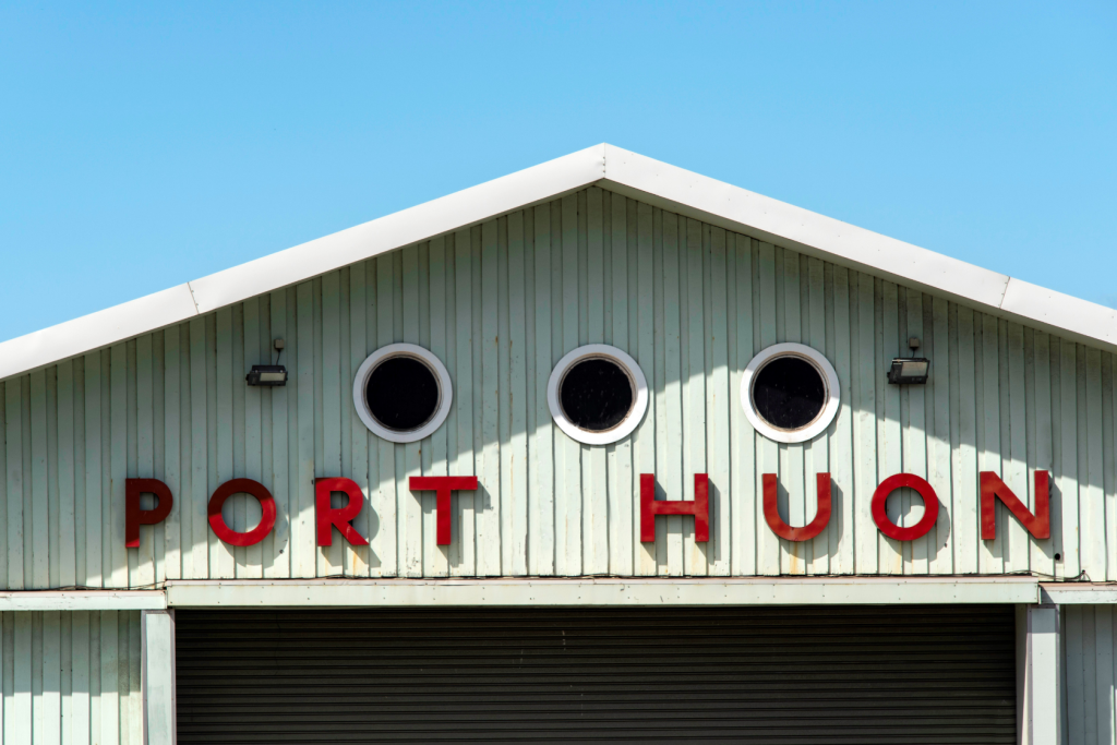 The upper facade of a pale green building with PORT HUON spelled in large red letters and three round windows, against a clear blue sky.