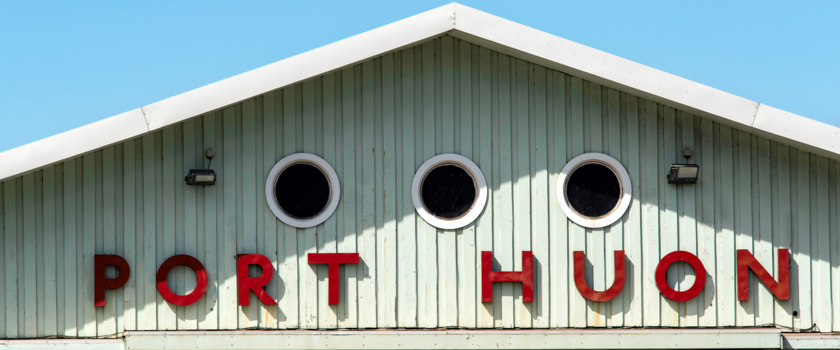 The upper facade of a pale green building with PORT HUON spelled in large red letters and three round windows, against a clear blue sky.