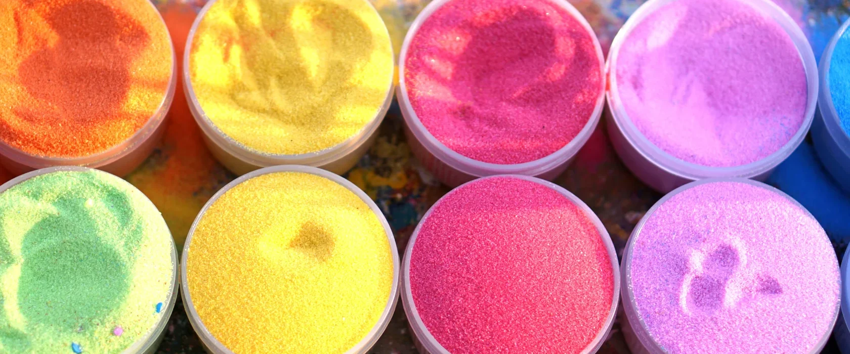 Eight containers filled with brightly colored sand—orange, yellow, pink, purple, green, and red—are arranged on a messy, paint-splattered surface. Some sand has finger impressions.