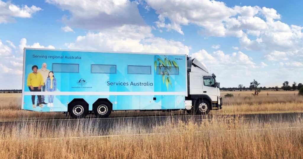 A large blue Services Australia truck drives along a rural road through dry grasslands under a partly cloudy sky, displaying images of people and the words “Serving regional Australia.”.