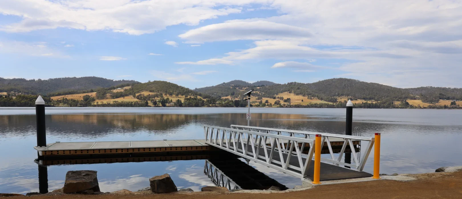 A metal dock with a ramp extends over a calm lake, surrounded by hills and trees under a partly cloudy sky. Reflections of the clouds and landscape are visible on the water’s surface.
