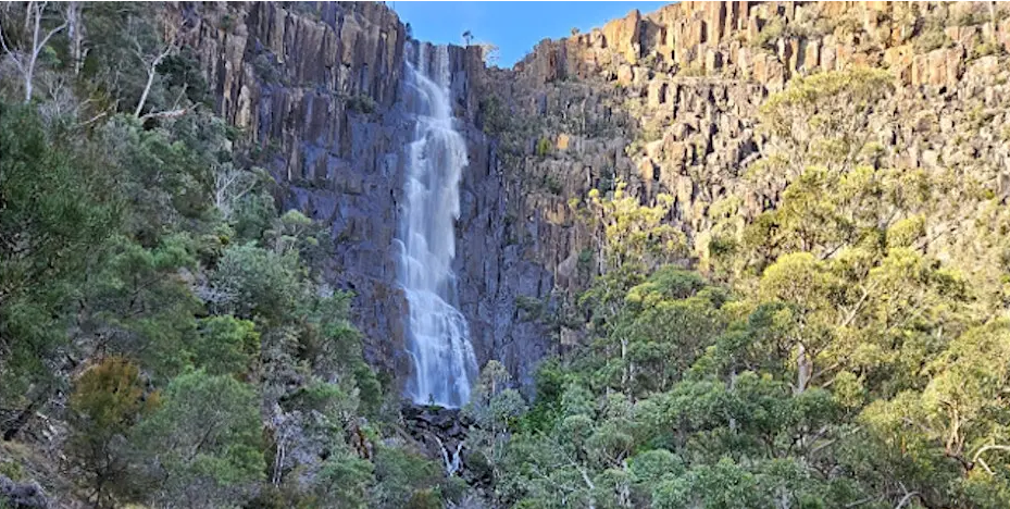 Tall waterfall cascading down a rocky cliff surrounded by lush green trees and vegetation under a clear blue sky.