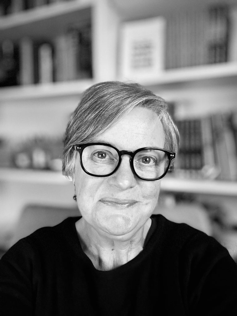 A person with short gray hair and black glasses smiles at the camera. They are wearing a dark top, and behind them are shelves filled with books. The image is in black and white.