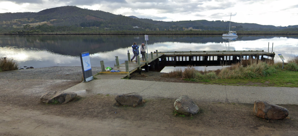 A wooden pier extends into a calm lake with two people standing at the end. A white sailboat is anchored on the water, and hills covered in trees rise in the background under a cloudy sky.
