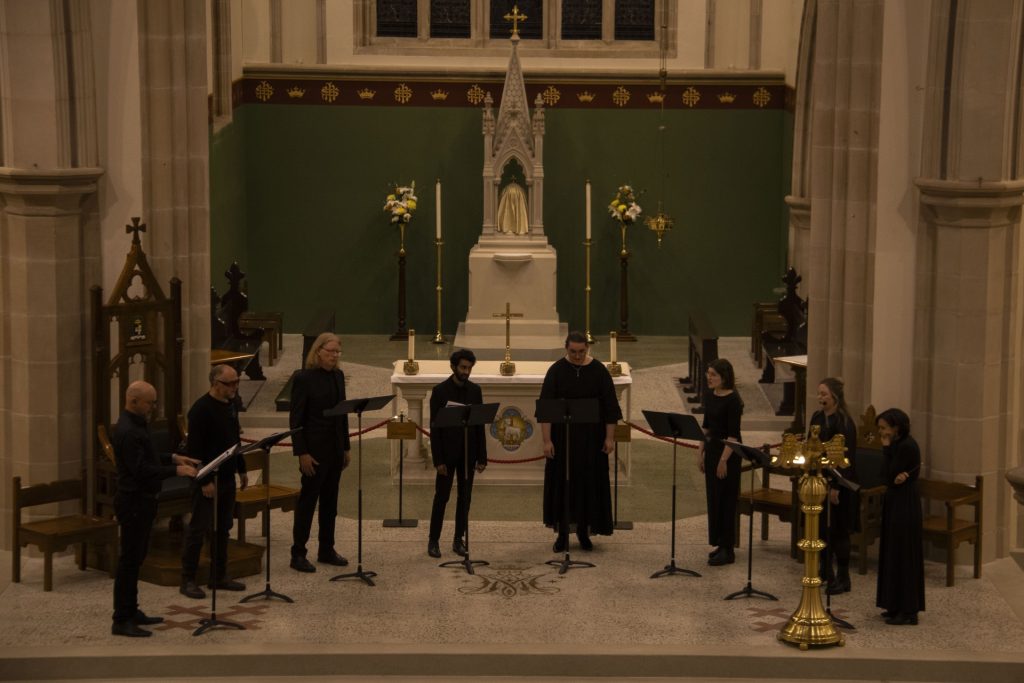 A group of eight people dressed in black sing from sheet music on stands in a church, standing in a semicircle before an altar with religious statues and candles.