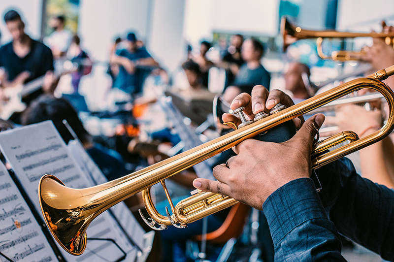 A close-up of a person playing a trumpet in an orchestra, with music sheets visible and other musicians, including a guitarist, blurred in the background.