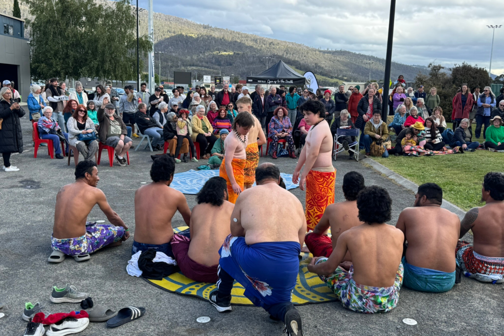 A group of men wearing colorful traditional skirts perform a cultural dance outdoors, encircled by a large seated and standing audience, with mountains and cloudy skies in the background.