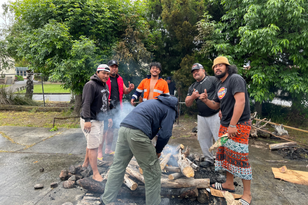 Six men stand around a smoky outdoor fire pit with logs, some smiling and giving thumbs up. Trees and a fence are in the background. One man is tending to the fire with his back to the camera.