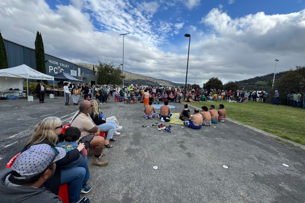 A diverse crowd sits and stands outdoors near a building, watching a group of dancers in traditional attire performing on a paved area under a partly cloudy sky. Hills and trees are visible in the background.