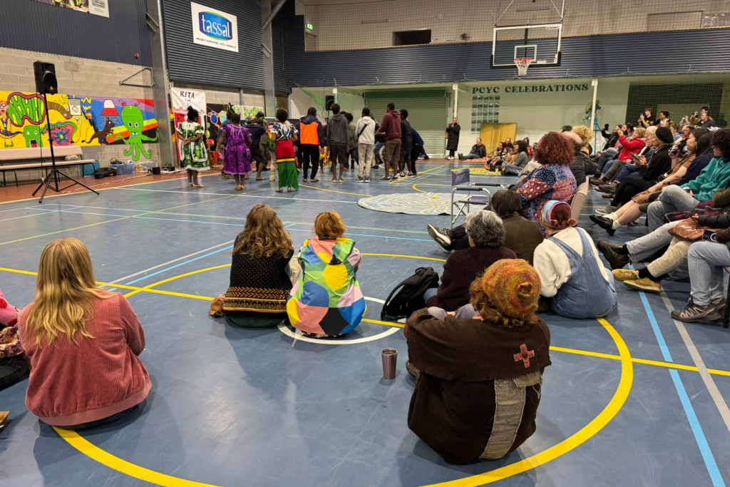 A diverse group of people sit on a gym floor, watching performers in colorful costumes on a stage. The gym has a basketball hoop, wall art, and a banner reading “PCYC Celebrations.”.