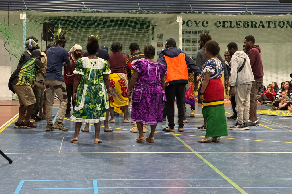 A group of people, some in colorful traditional clothing, dance in a gymnasium while others watch from the side. There is a net in the background and the words PCYC CELEBRATION on the wall.