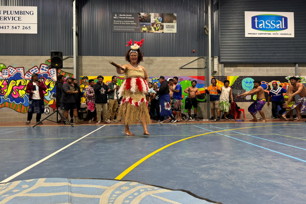 A performer in traditional Pacific Island attire dances in a gymnasium, while a group of people in colorful outfits watch from the background. The walls feature murals and company signs.