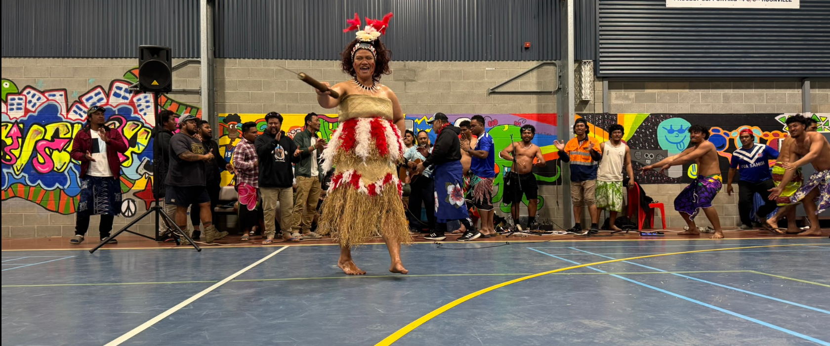 A performer in traditional Pacific Island attire dances in a gymnasium, while a group of people in colorful outfits watch from the background. The walls feature murals and company signs.