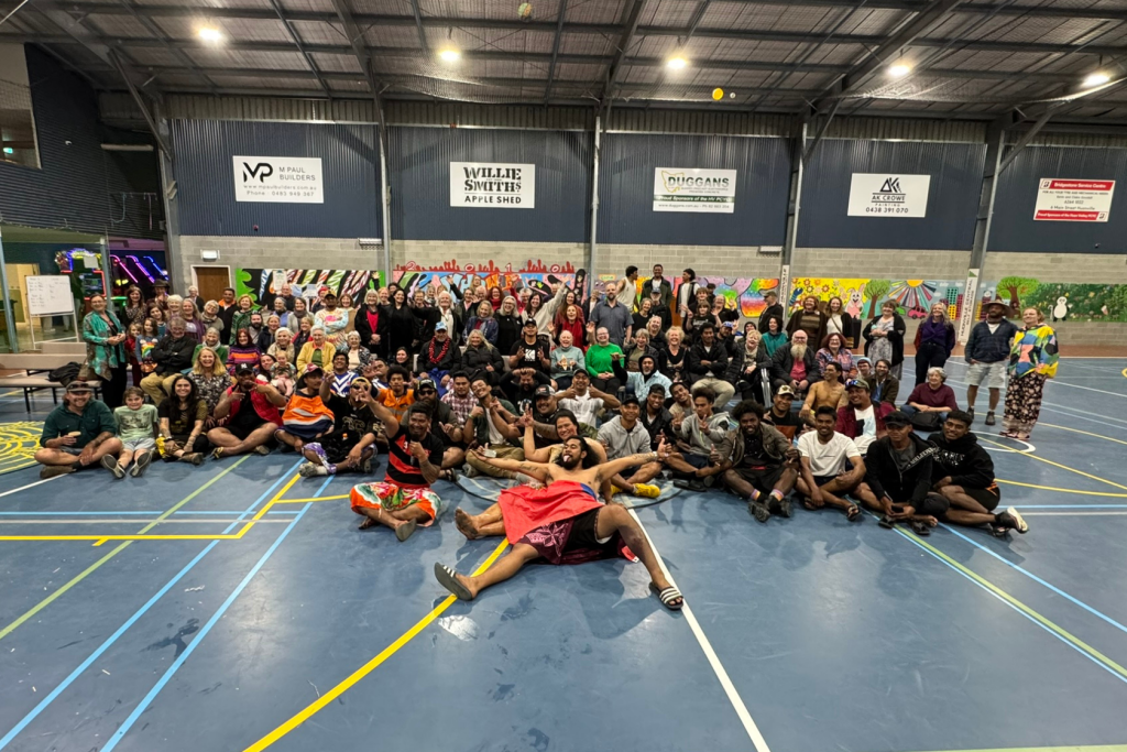 A large, diverse group of people, including teens and adults, pose together in a gymnasium with colorful murals on the walls. Some are sitting cross-legged on the floor, while others stand behind them, smiling at the camera.