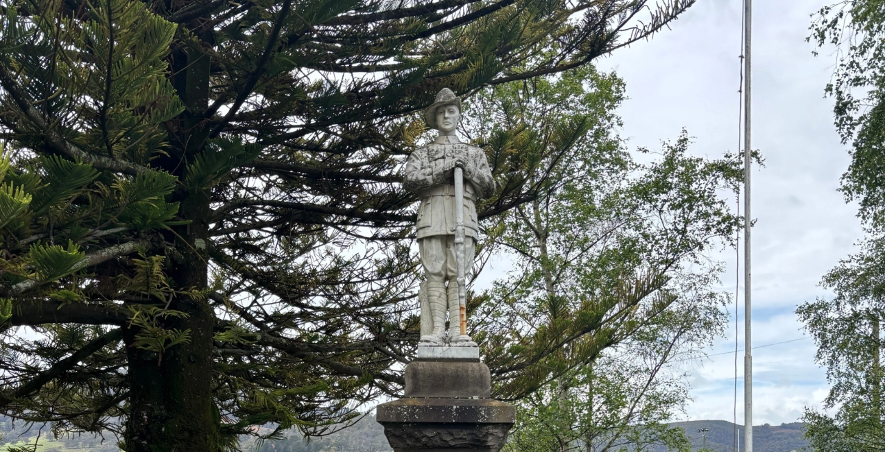 A stone statue of a soldier stands on a pedestal in an outdoor setting, surrounded by green trees, with a cloudy sky and distant hills in the background.