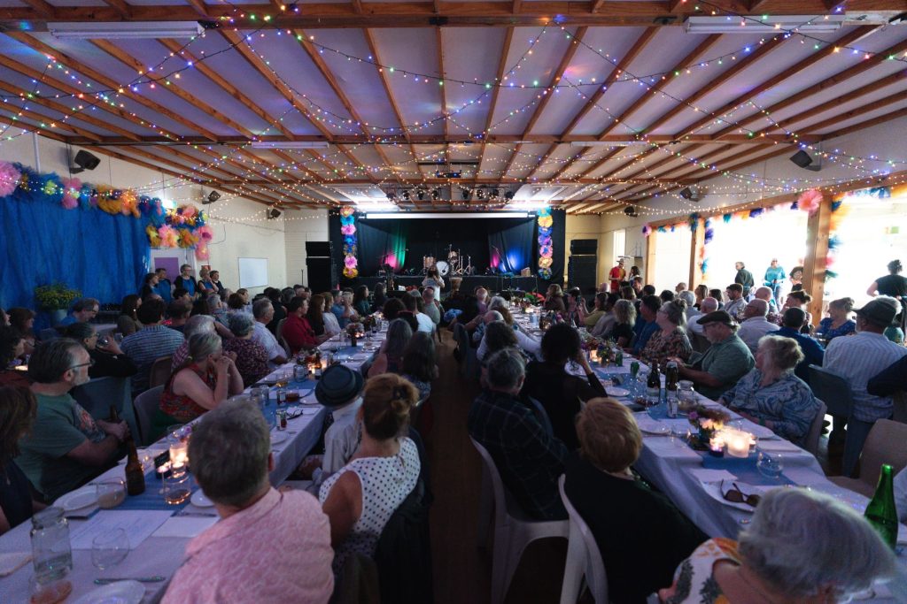 A large group of people sit at long tables decorated with candles and flowers in a festively lit hall, watching a performance on stage under colorful string lights and vibrant decorations.