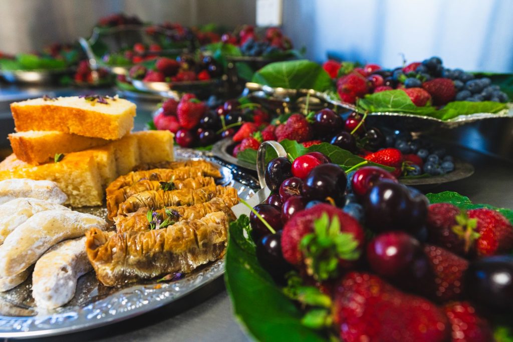 A close-up of assorted desserts and fresh fruits on silver trays, including cakes, pastries, powdered sugar cookies, and a variety of berries and cherries, all arranged on green leaves.