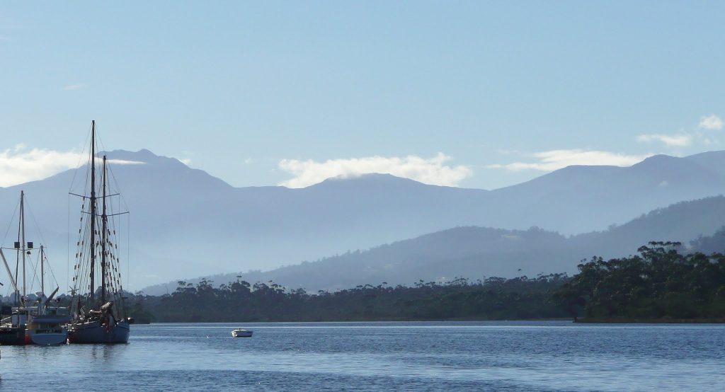 A calm body of water with sailboats docked on the left, lush green shoreline, and misty mountains in the background under a clear blue sky.