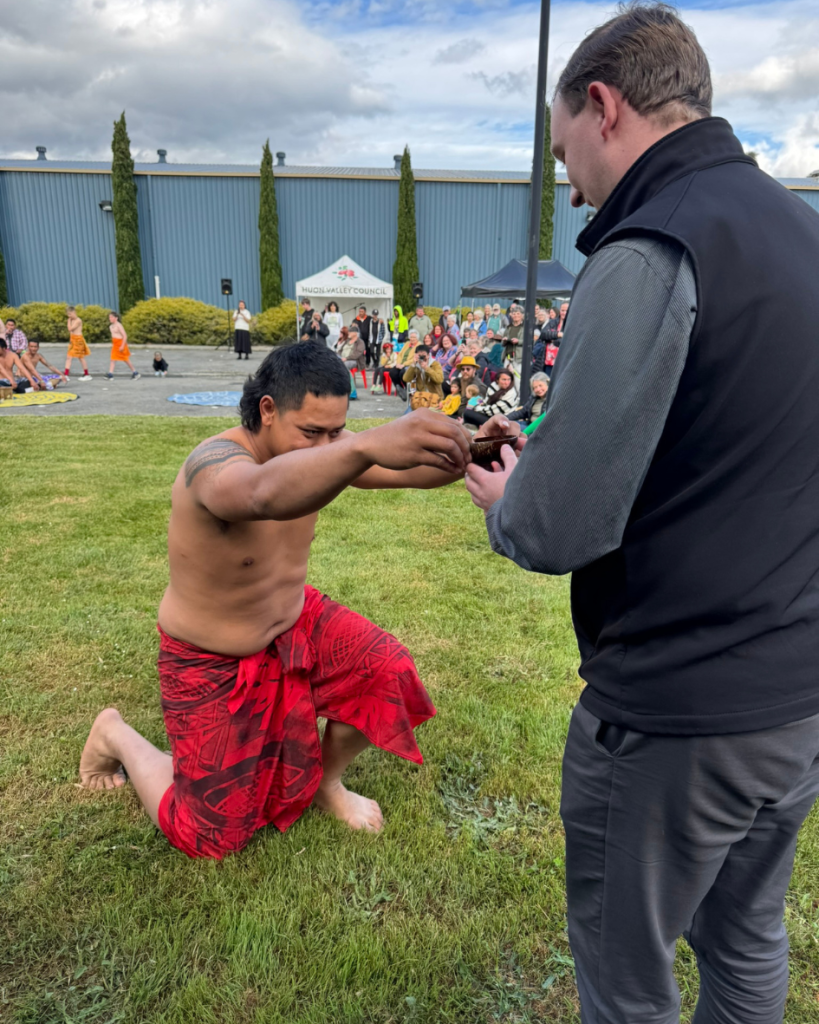 A man in a red sarong kneels on grass, offering an object to another man standing in front of him. A crowd watches in the background, with tents and a blue building visible.