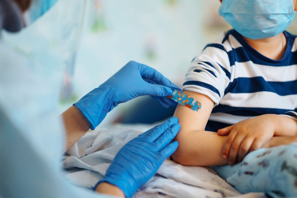 A healthcare worker in blue gloves places a colorful adhesive bandage on a child’s arm after vaccination. The child, wearing a striped shirt and face mask, sits with their other arm crossed.