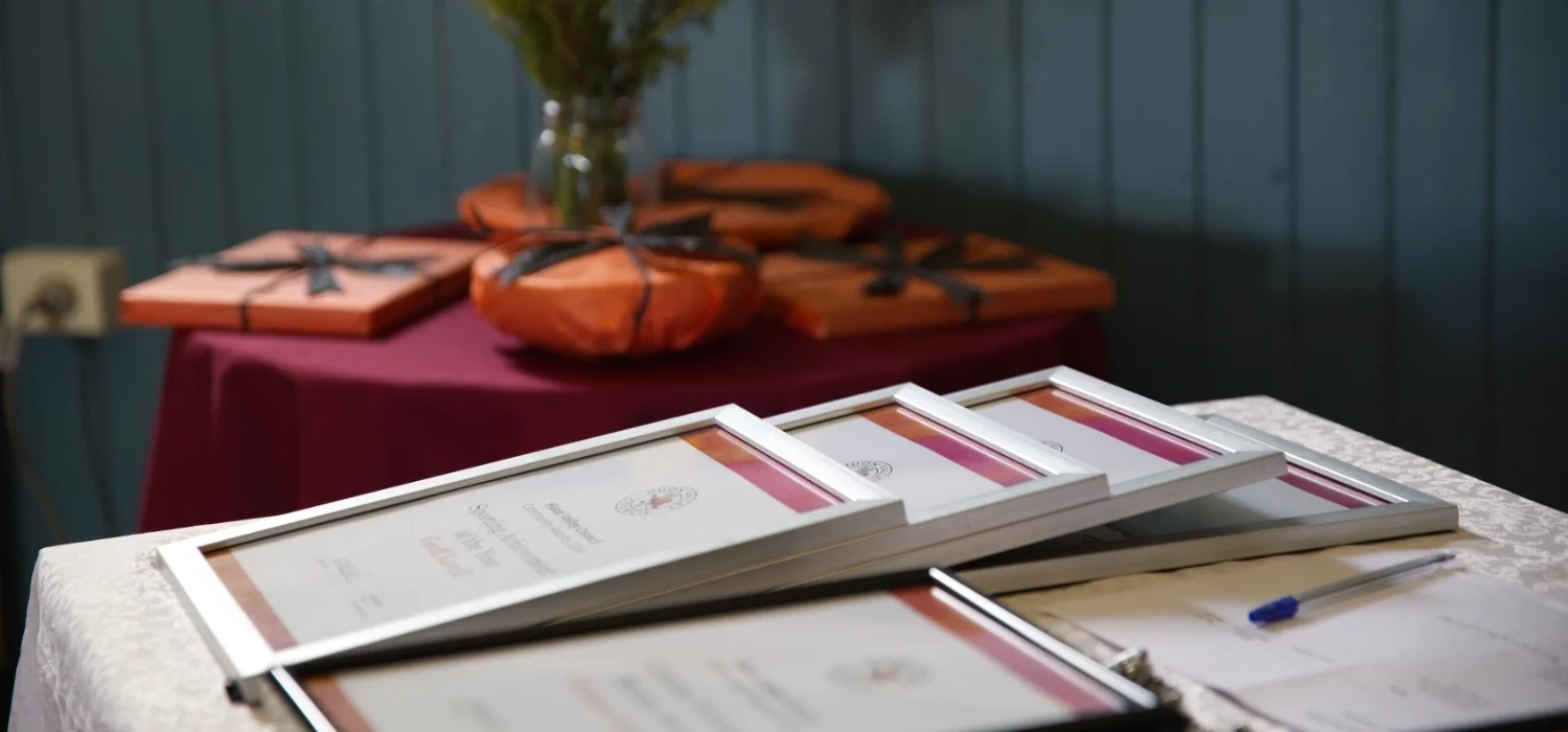 Several framed certificates are displayed on a table covered with a white cloth. In the background, wrapped gifts with black ribbons and a vase of yellow flowers sit on a round table with a maroon tablecloth.
