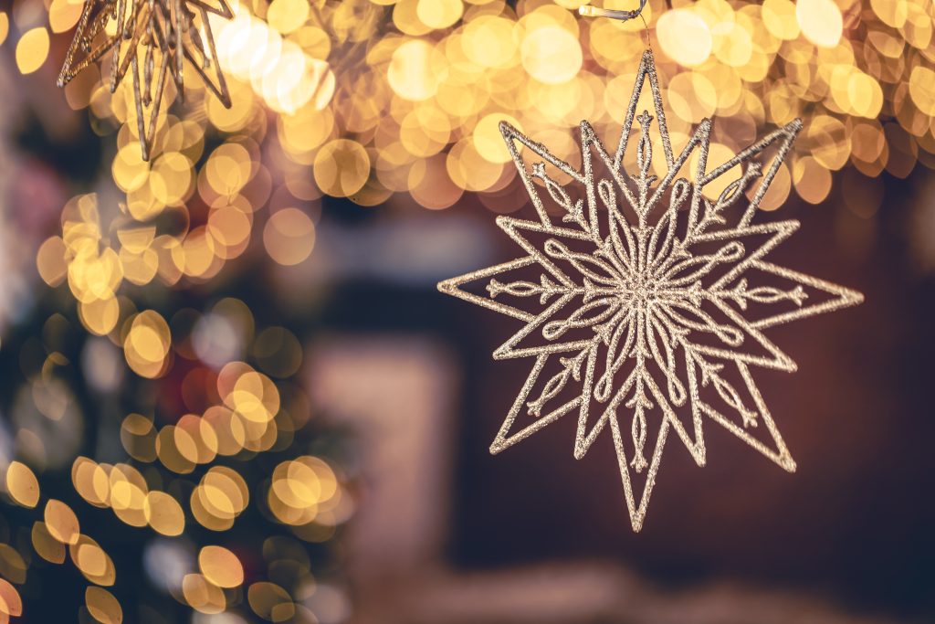 A glittery snowflake ornament hangs in focus, with blurred golden holiday lights and a Christmas tree in the background, creating a festive and warm atmosphere.