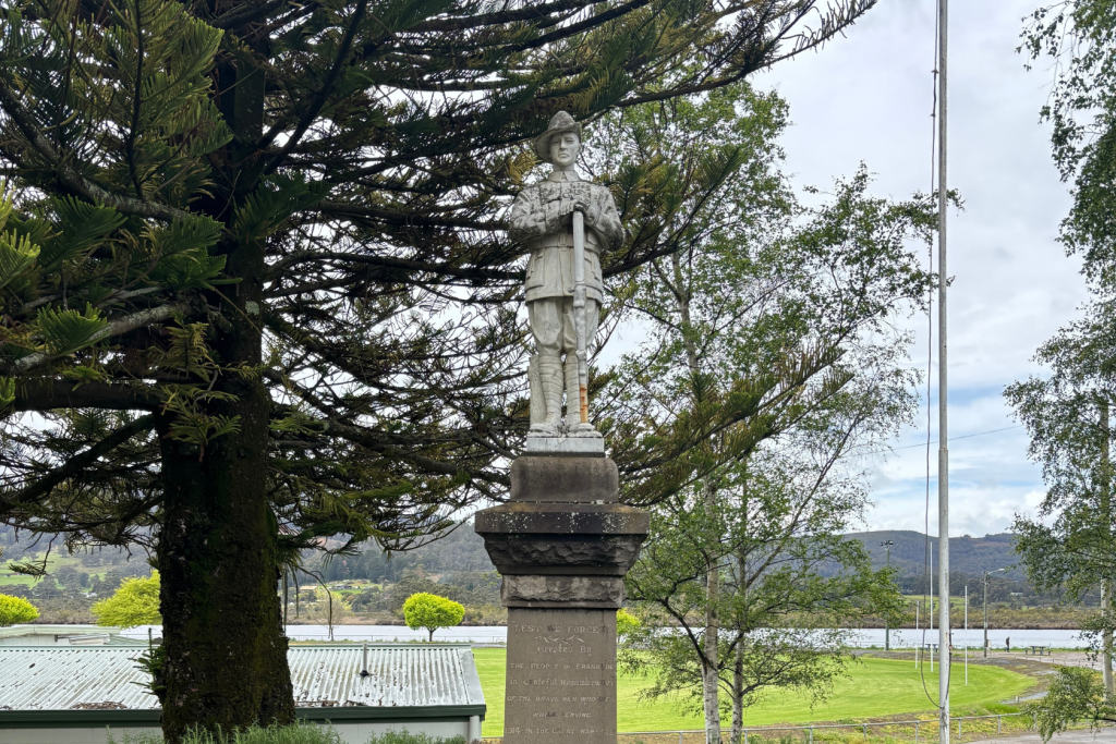 A stone statue of a soldier holding a rifle stands on a pedestal surrounded by trees. In the background are a field, a shed, and a river with hills under a cloudy sky.