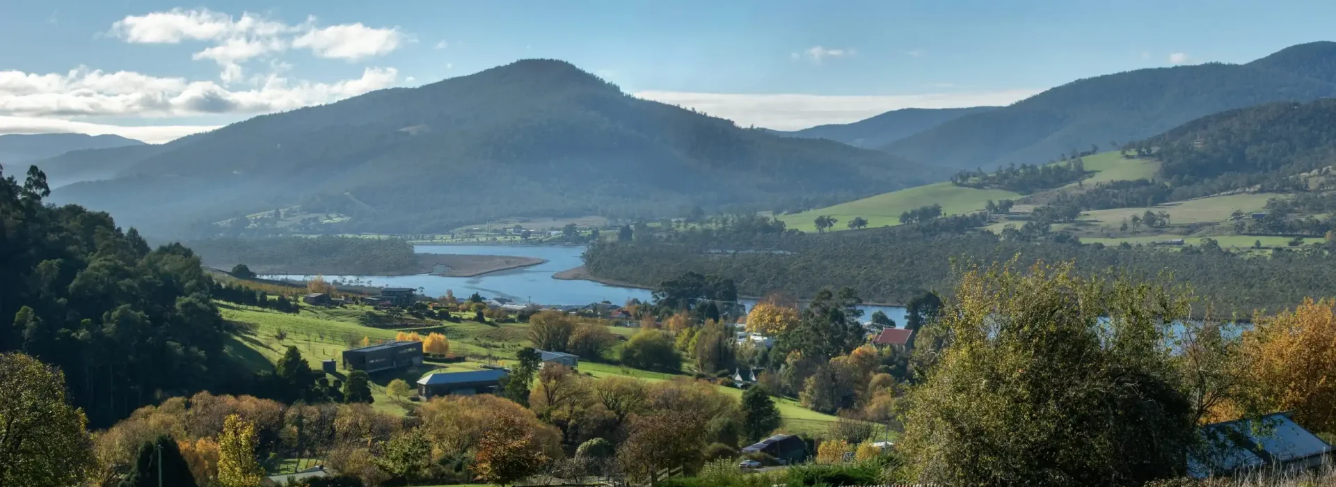 A scenic rural landscape with rolling green hills, scattered houses, trees with autumn foliage, a winding river, and mountains in the background under a partly cloudy sky.