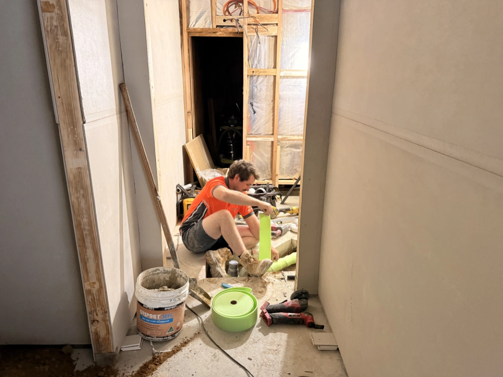 A person in an orange shirt and shorts sits on the floor of a construction site, working with green pipes amid tools, buckets, and building materials in a hallway with unfinished walls.