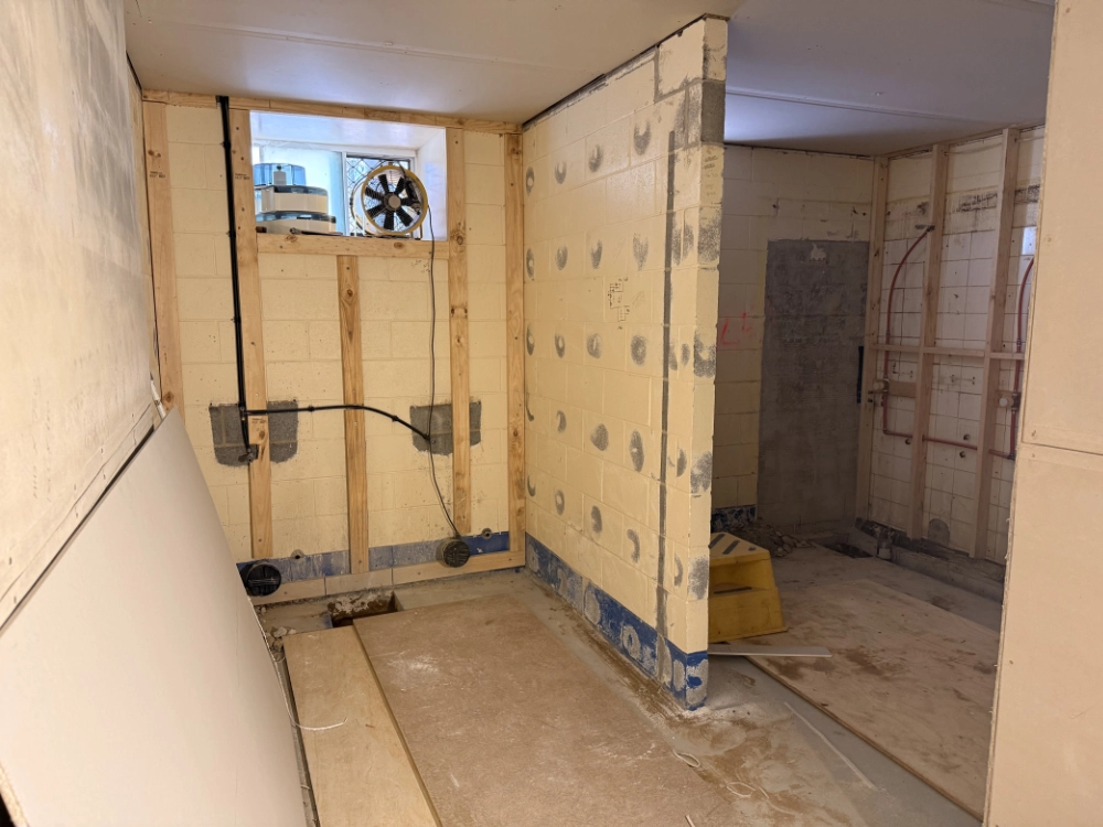 A room under construction with exposed masonry walls, wood framing, and visible wiring. There is a small window with a fan, boards leaning on a wall, and construction materials scattered on the dusty floor.