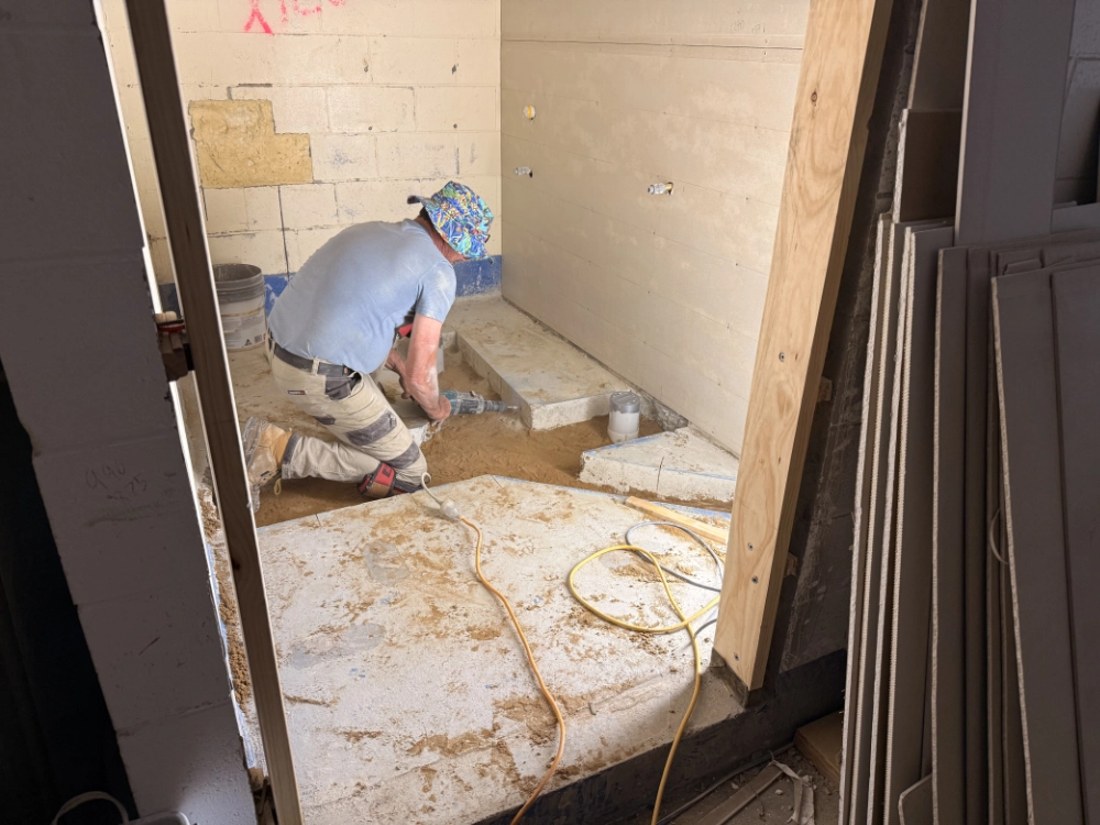 A construction worker wearing a hat and gloves is kneeling on the floor, working with tools and sand inside an unfinished room with exposed brick and drywall panels leaning against the wall.