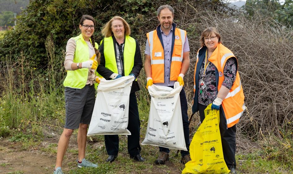 Four adults in safety vests stand outdoors, smiling and holding bags filled with collected litter. They are participating in a community clean-up event near bushes and grass, with trees and hills visible in the background.