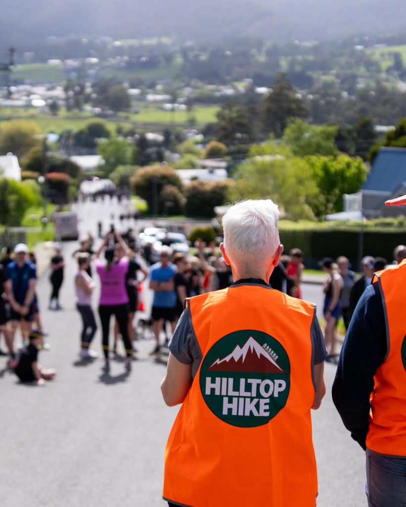 Two people in bright orange vests with Hilltop Hike logos stand facing a crowd of hikers gathered on a sunny, tree-lined street with a scenic valley and hills in the background.