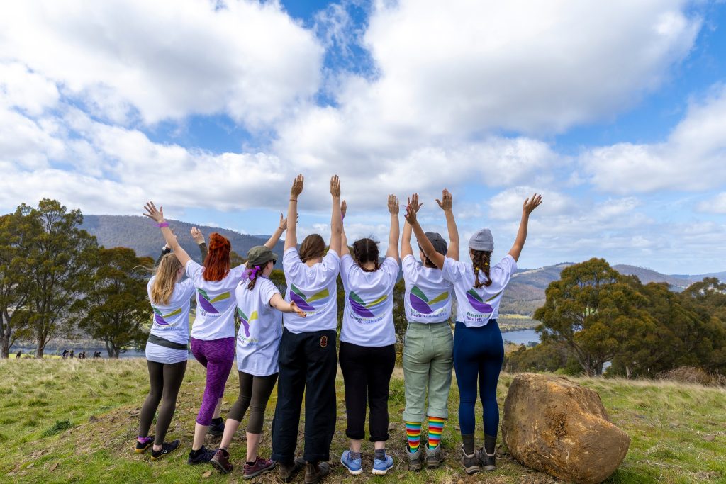 A group of seven people stand outdoors on grass with their backs to the camera, arms raised joyfully. They wear matching white T-shirts and face a scenic landscape of hills, trees, and a partly cloudy sky.