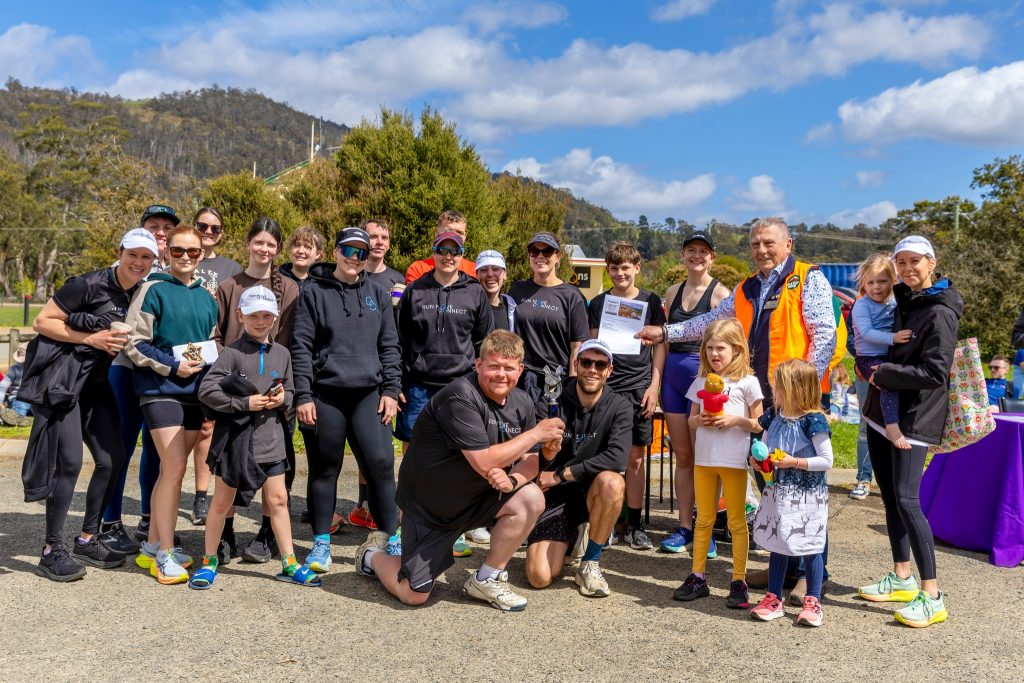 A large group of people, including adults and children, pose outdoors in sportswear on a sunny day. Some hold certificates and trophies, and there are hills and trees in the background.