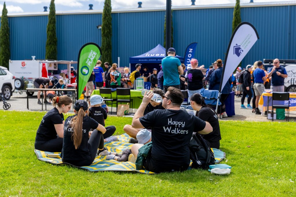 A group wearing Happy Walkers shirts sits on a picnic rug on grass, facing away, at an outdoor event with tents, banners, and other people in the background. One person is holding a water bottle.