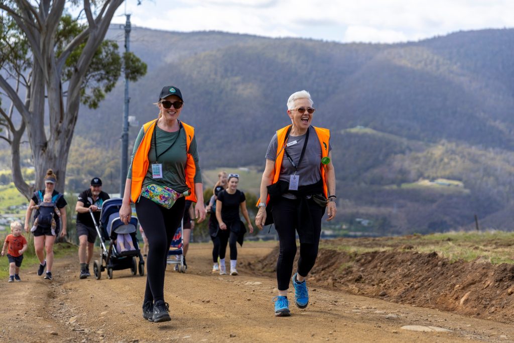 A group of people walks outdoors on a dirt path in a hilly, rural area. Two women in orange safety vests lead, smiling, while others follow behind, including children and someone pushing a stroller.