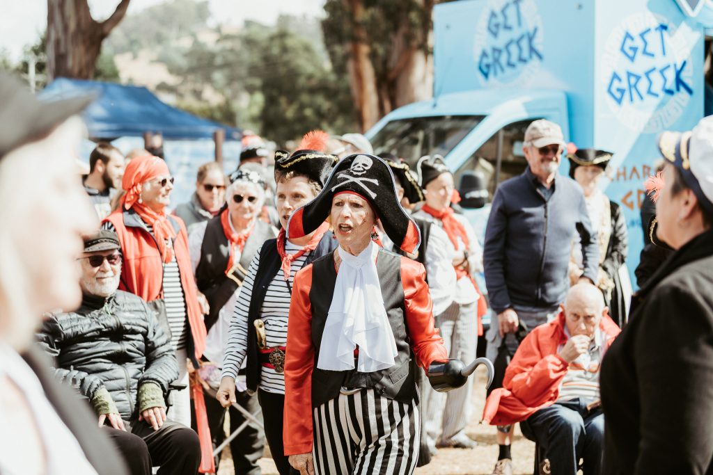 A group of adults dressed as pirates gather outdoors at an event. One woman in a pirate hat, red jacket, and striped pants stands out in the center, while others sit or stand around her. A blue food truck is in the background.