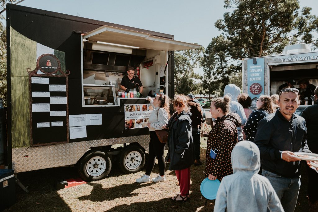 People wait in line at a food truck during an outdoor event. The vendor inside serves food while customers, some holding plates, stand on grass under sunny skies with trees in the background.