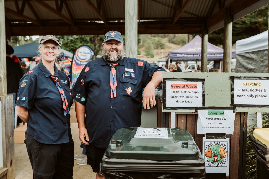 Two adults in scout uniforms stand and smile beside waste, recycling, and compost bins at an outdoor event. Signs detail waste separation, and a covered area with people and stalls is visible in the background.