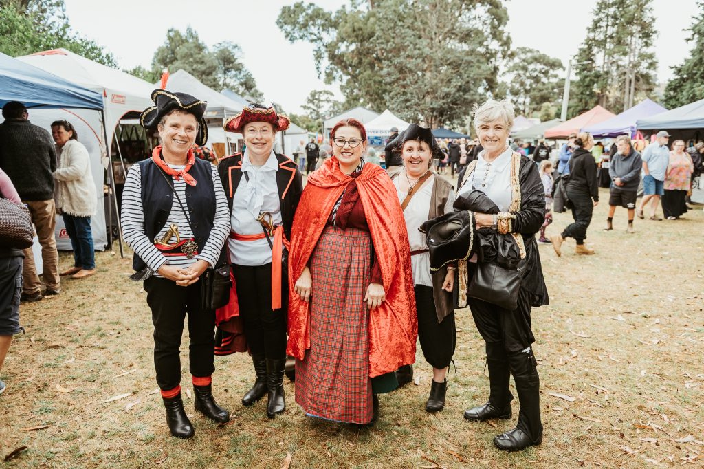 Five people in colorful, historical costumes pose and smile at an outdoor festival with tents and trees in the background. The group includes pirates and a woman in a red cape and tartan skirt.