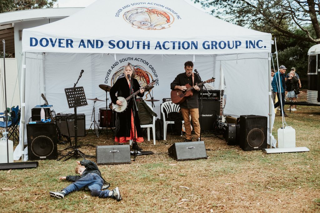 Musicians perform under a white tent labeled Dover and South Action Group Inc. while a child lies playfully on the grass in front of the stage at an outdoor event.