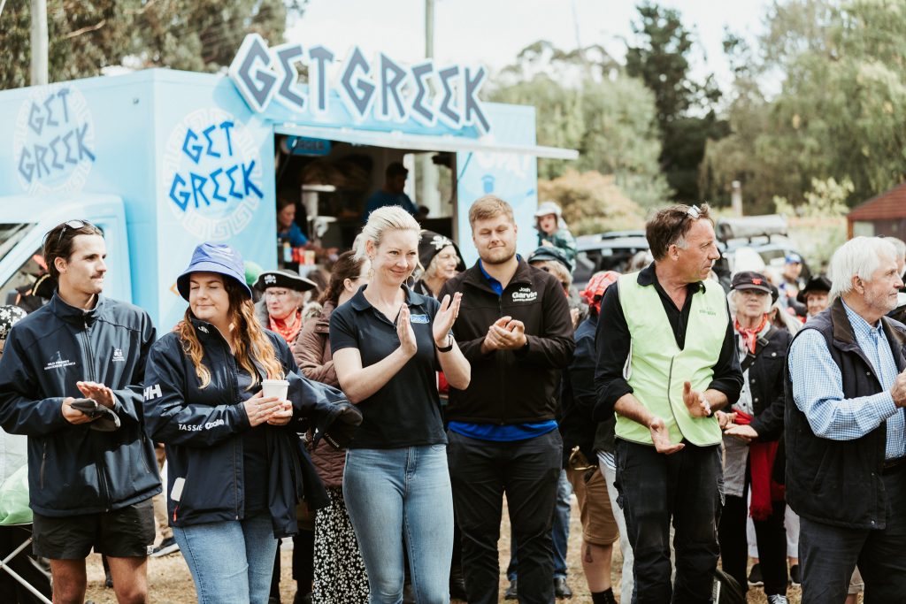 A group of people stands outdoors, clapping and smiling in front of a blue food truck labeled “GET GREEK.” Some wear jackets and hats, and trees are visible in the background.