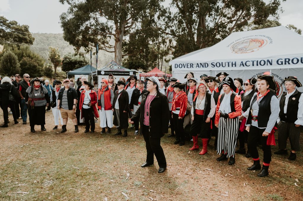 A large group of people in pirate costumes stands on grass near tents at an outdoor event, beneath trees and a cloudy sky. The tents display the name “Dover and South Action Group.”.