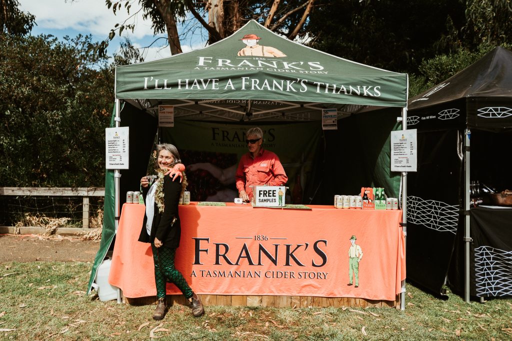 A smiling woman stands with a drink in front of a Frank’s cider stall at an outdoor event, where a person behind the counter offers free samples. The stall is green with orange branding and surrounded by trees.