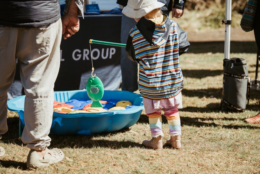 A child in colorful clothes holds a toy fishing rod, catching a green plastic fish from a small blue kiddie pool, with adults standing nearby outdoors.