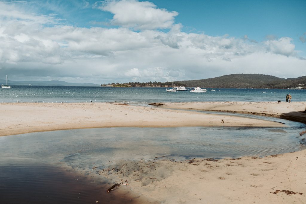 A sandy beach with a small stream flowing into the sea, sailboats anchored in the blue water, and distant hills under a partly cloudy sky. A person walks along the shore to the right.