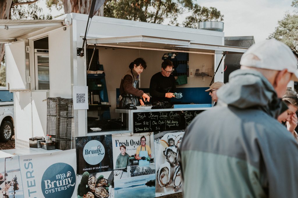 Two people prepare and serve oysters inside a white food truck decorated with oyster posters and menus, while customers wait outside. Trees and other market stalls are visible in the background.